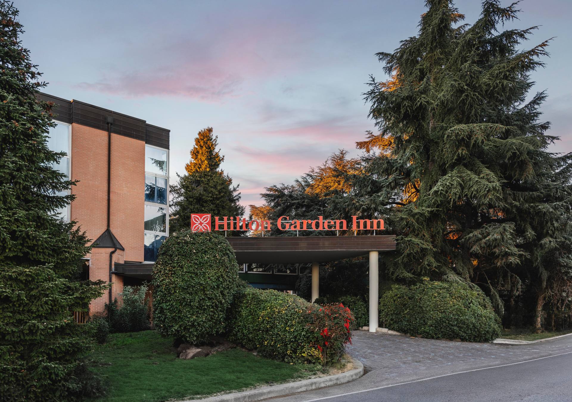 Hilton Garden Inn Bologna North Exterior, signage, trees, bushes, driveway, at sunset