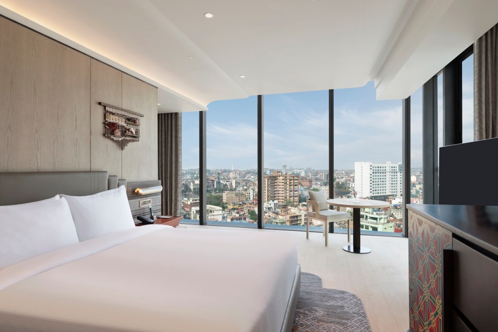 Hilton Kathmandu - King Room with Balcony, floor to ceiling windows, view of the city, curtain, chair, table, side table, phone TV, chest of drawers, bed