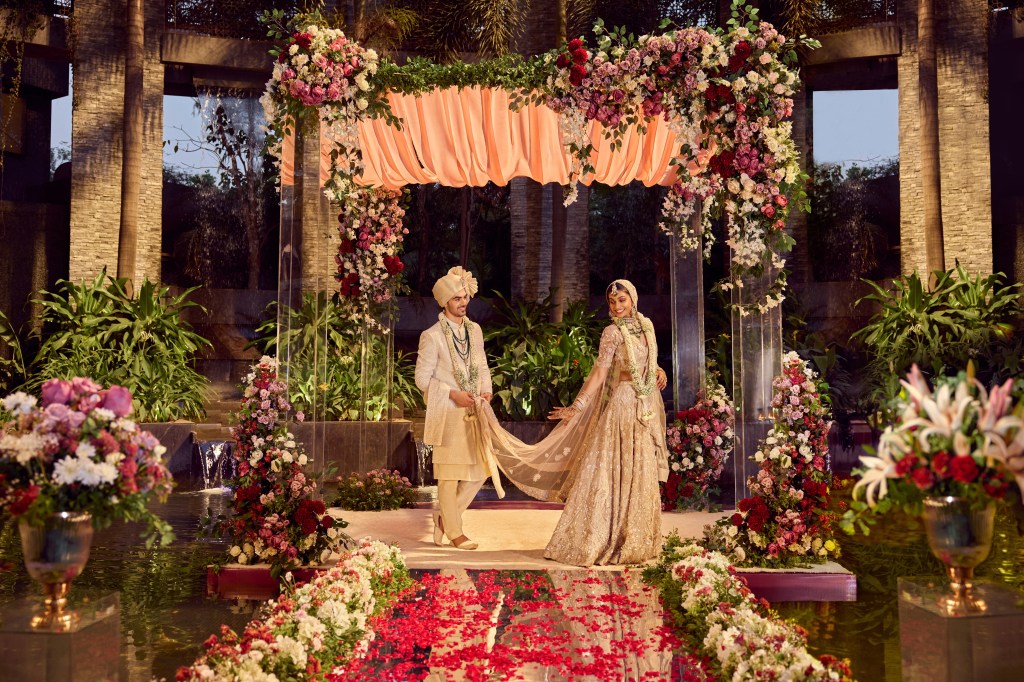 man and woman at wedding alter in Indian wedding attire, flower arch, flowers in the aisles,rose petals on mirrored walkway