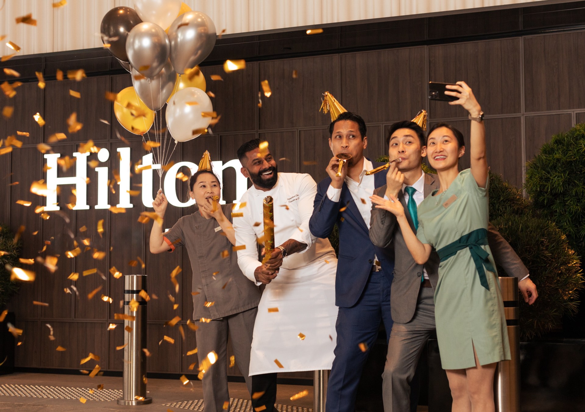 A group of five Hilton Team Members celebrate and take a selfie with balloons, party hats and confetti in front of a Hilton sign