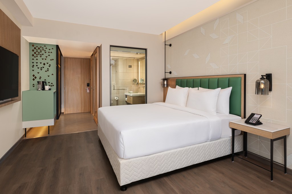 Hilton Garden Inn Surat City Centre, King Guest Room, green headboard, white bedding, green dry bar, white night stand table, glass sconce lamp, white pillows, television mounted on wall, wood floors, room entrance, bathroom, interior window wall