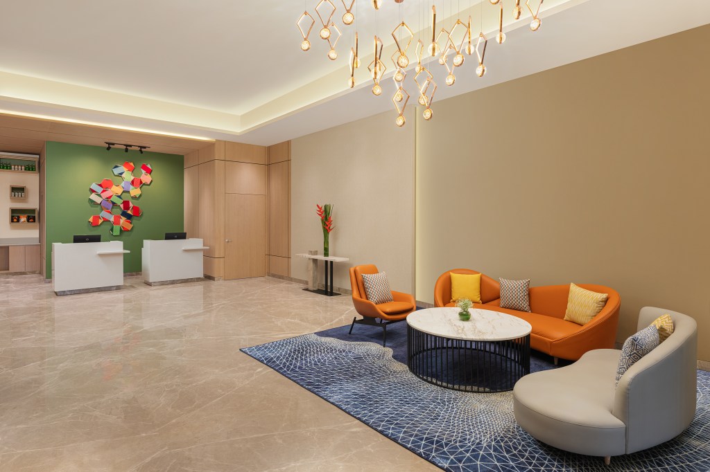 Hilton Garden Inn Surat City Centre, Lobby, orange lounge furniture, orange couch, orange accent chair, hanging artwork, modern ceiling light fixture, marble topped black round coffee table, blue designed area rug