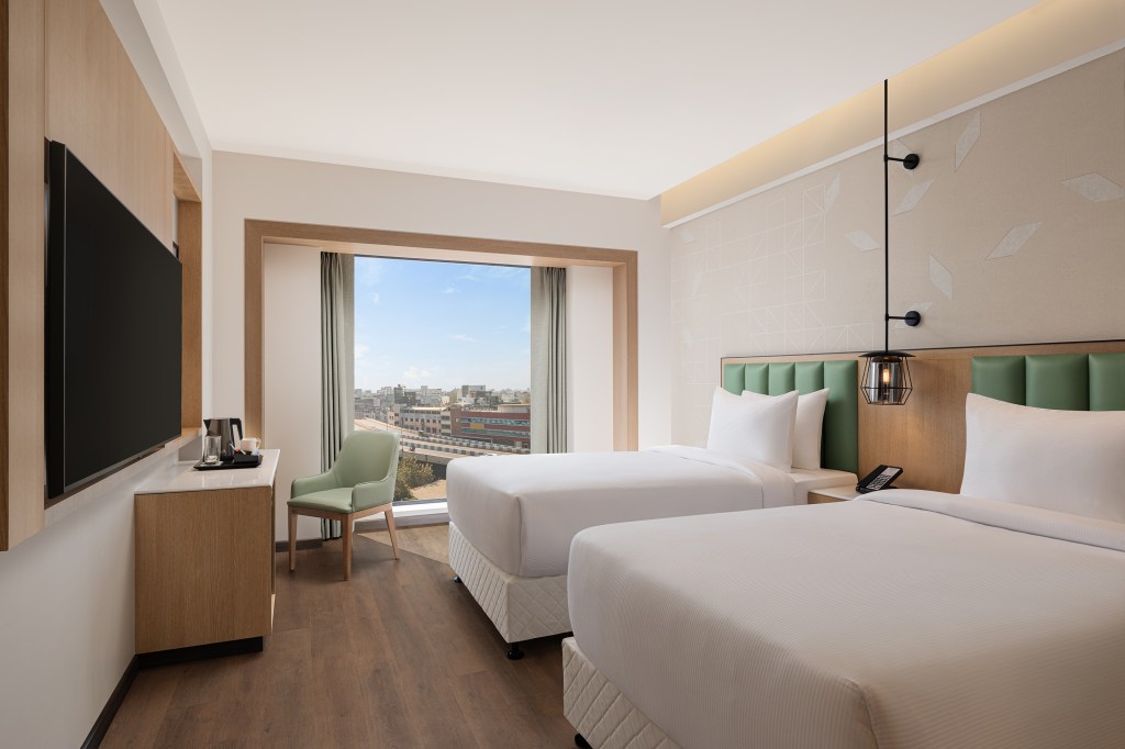 Hilton Garden Inn Surat City Centre, Twin Deluxe City View, green headboard, green chair, black sconce light, mounted television, wooden floors, curtains, large window, white bedding, white pillows