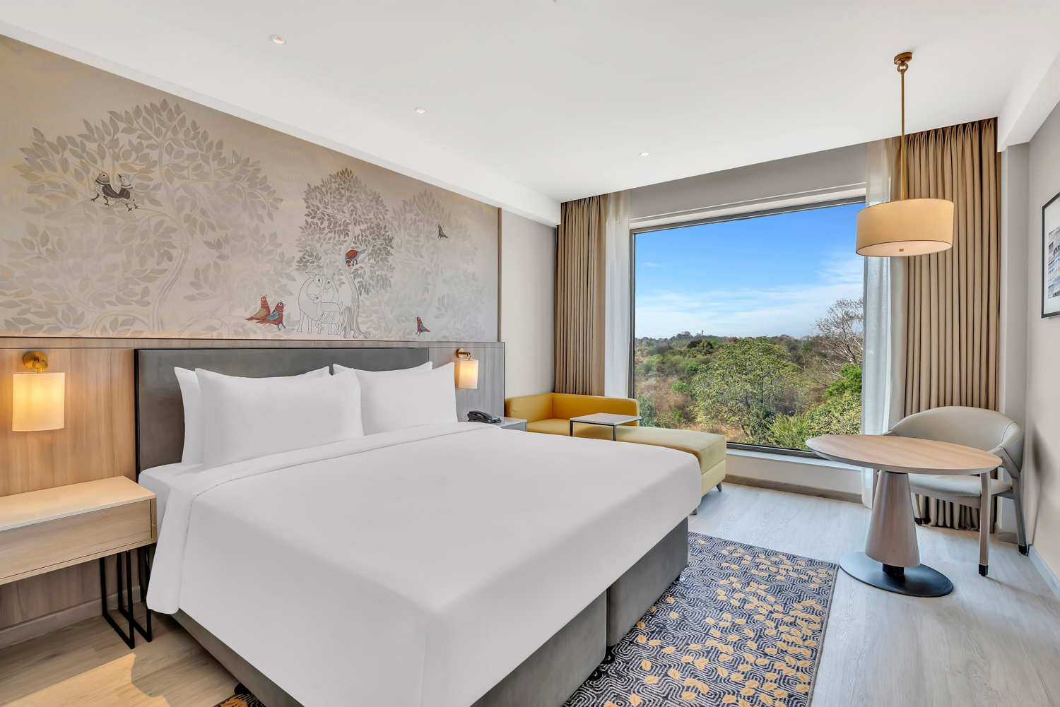 King Guest Room with white bedding and side tables with a phone on it and a sconce light, Chaise lounge couch seating area with a window view, sitting area with a pendant light above and hanging framed art