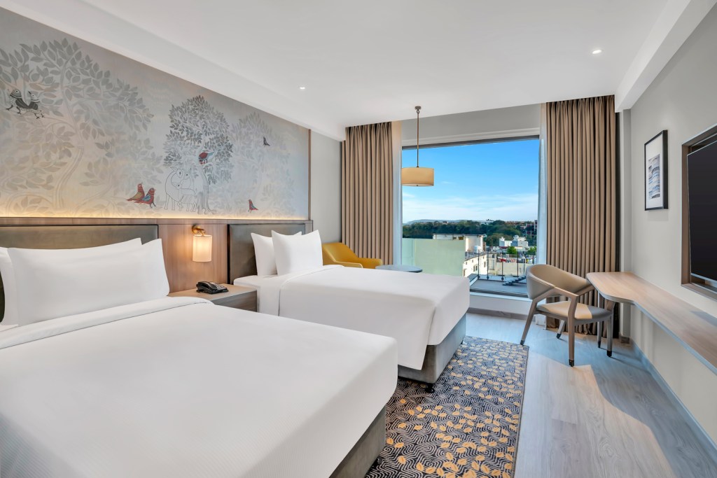 Bedroom with two twin beds with white bedding and a side table with wall sconce light and a phone on it, yellow chair with a window view with a pendant light above and a sitting area with a framed picture