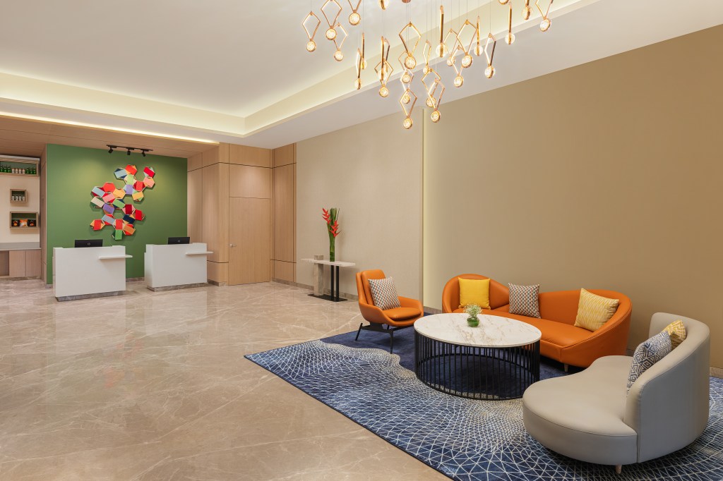 Hilton Garden Inn Surat City Centre rendering, lobby and reception area with colorful orange and cream seating, blue rug, modern lighting fixture, green wall with colorful artwork behind check in desk