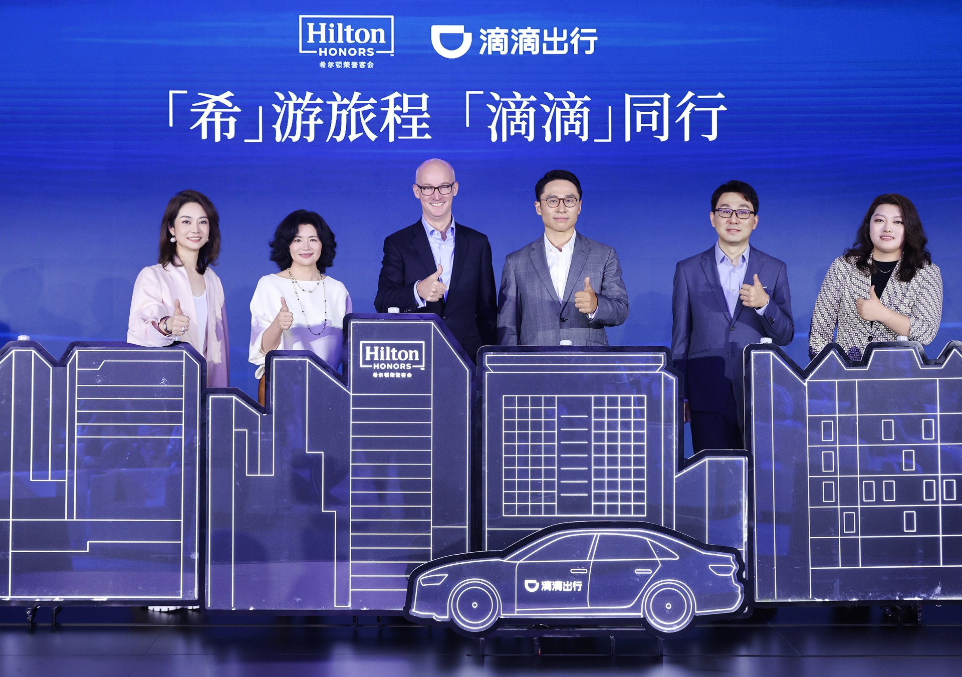 Hilton Honors and DiDi ChuXing Announce Partnership - group of 6 people pose for photo with Hilton Honors and DiDi ChuXing logos behind a small blue cityscape with a car 希尔顿集团携手滴滴出行,共创住行一体会员体验新生态发布会