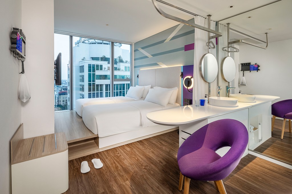 Twin Room with a window facing a city view that has two twin beds next to each other on a platform bed frame with wood flooring throughout and a purple accent chair at a counter facing a bathroom vanity with a sink
