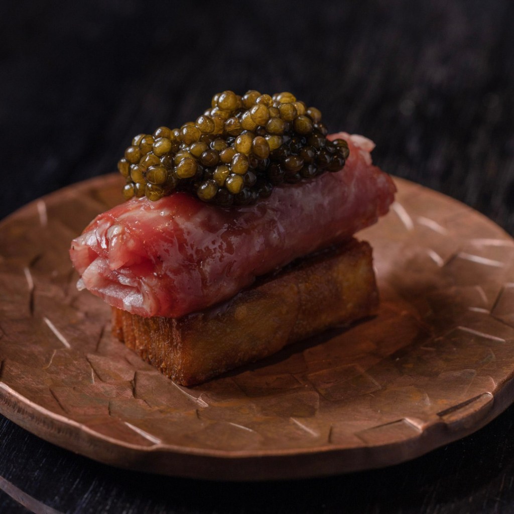 A decadent serving of steak tartare topped with a generous spoonful of glossy black caviar, served on a crispy rectangular potato base on a textured bronze plate.