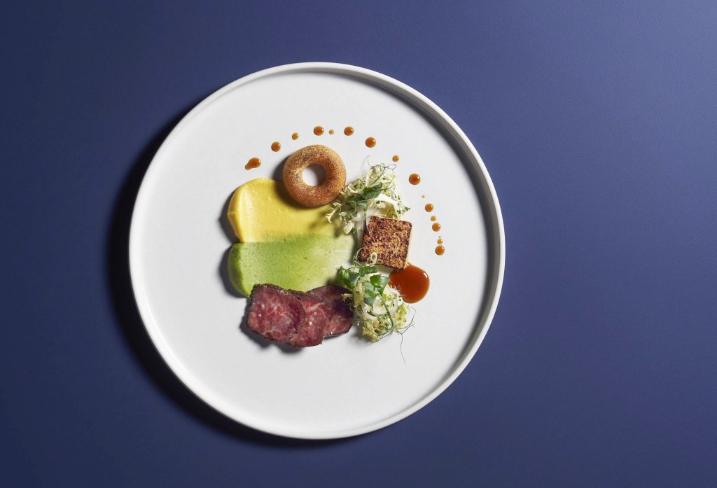 An artfully plated dish featuring seared Blackmore Wagyu slices, vibrant yellow and green purées, a small golden-brown pastry ring, salad greens, and a cube of seared meat, drizzled with sauce.