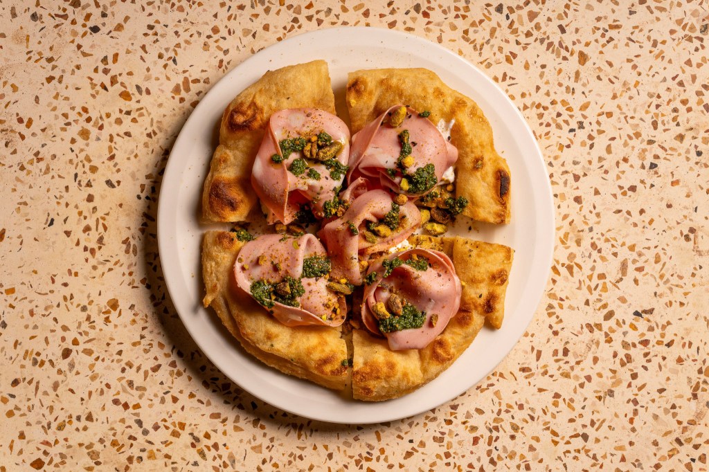 A plate of quartered fluffy focaccia topped with folds of mortadella, chopped pistachios, and drizzles of green herb sauce, served on a white plate.