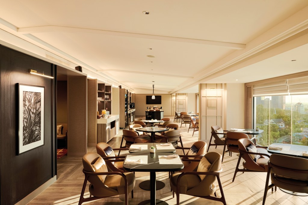 Hilton Kuching Executive Lounge dining sitting area with brown leather chairs at tables some next to windows that cover from floor to ceiling with a view of the city outside others next to a range from black walls with framed artwork to beverage stations
