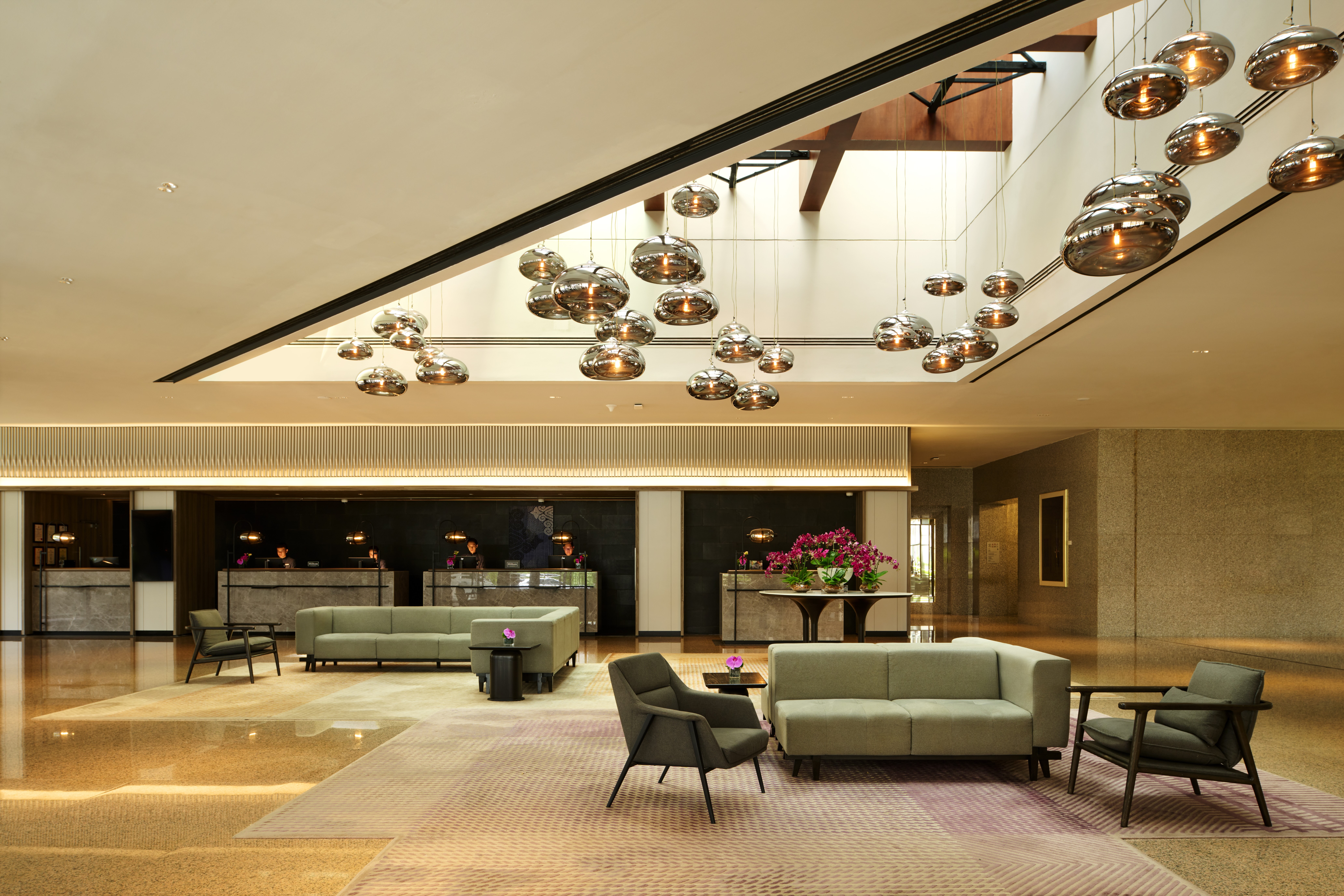 Hilton Kuching Lobby with two separate lounge seating areas with couches and end tables and lounge chairs next to high top tables with potted flowers with a cluster of strung pendant lights hanging from a skylight
