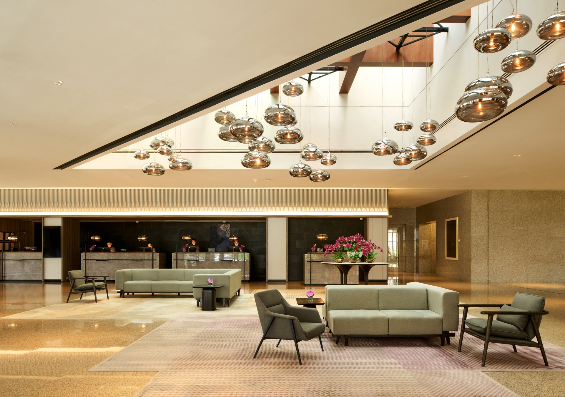 Hilton Kuching Lobby with two separate lounge seating areas with couches and end tables and lounge chairs next to high top tables with potted flowers with a cluster of strung pendant lights hanging from a skylight