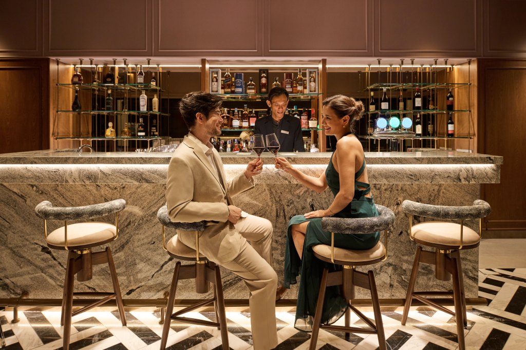 Hilton Kuching RYE bar where two people toast with red wine in stemmed glasses at a bar with black and white marble tiled floors and a marble bar top while they site on bar stools with wooden bases and a rounded back rest