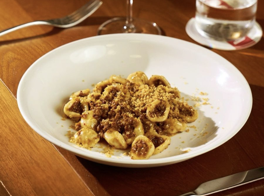 A white plate of orecchiette pasta coated in rich meat sauce and topped with golden breadcrumbs, paired with a glass of white wine and a glass of water on a wooden restaurant table.