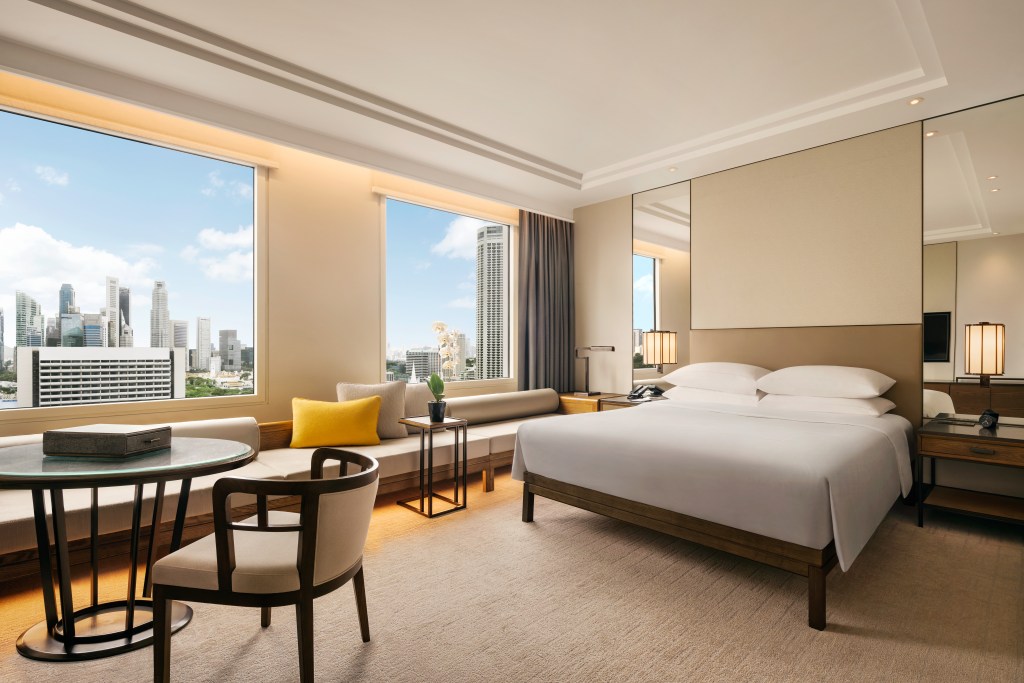 A room at Conrad Singapore Marina Bay with bed, table with chairs, and long bench seat under the large windows overlooking the city.