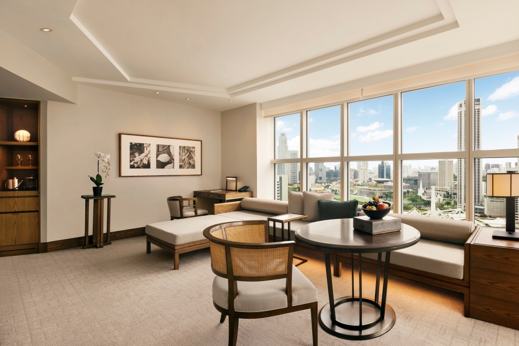 A room at Conrad Singapore Marina Bay featuring table with chair, and a long bench seat with chaise under the large windows overlooking the city.
