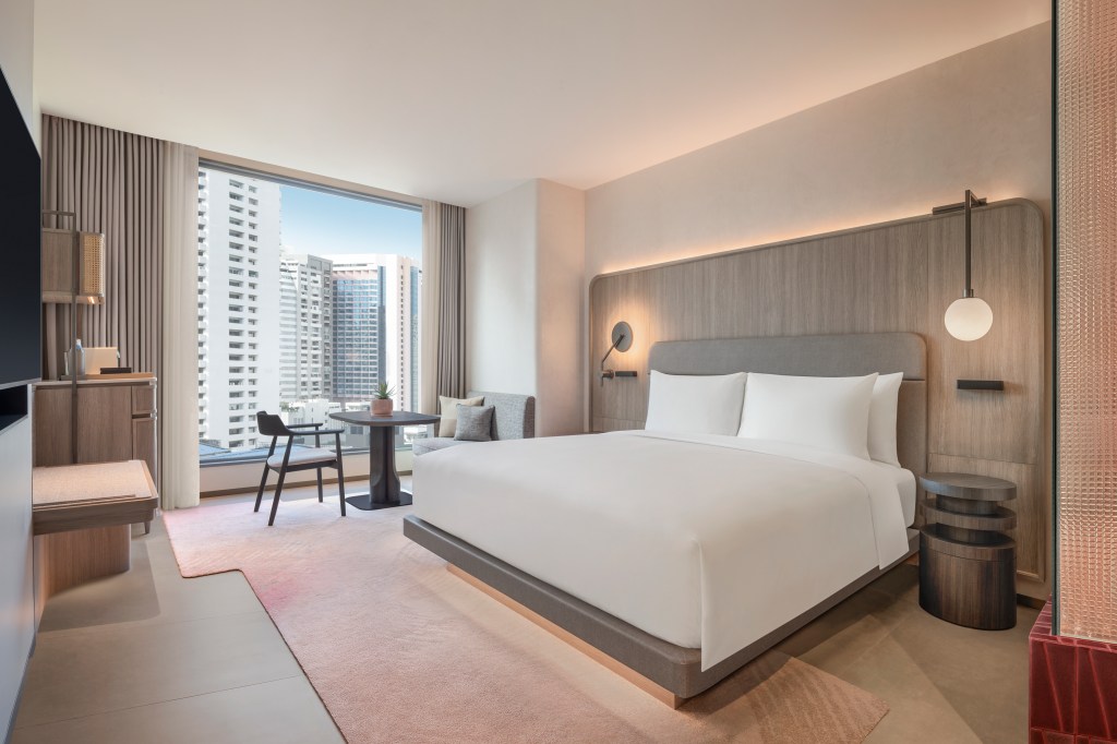 A bedroom with king bed, small tables and chairs, dresser, tv, and window overlooking the city.