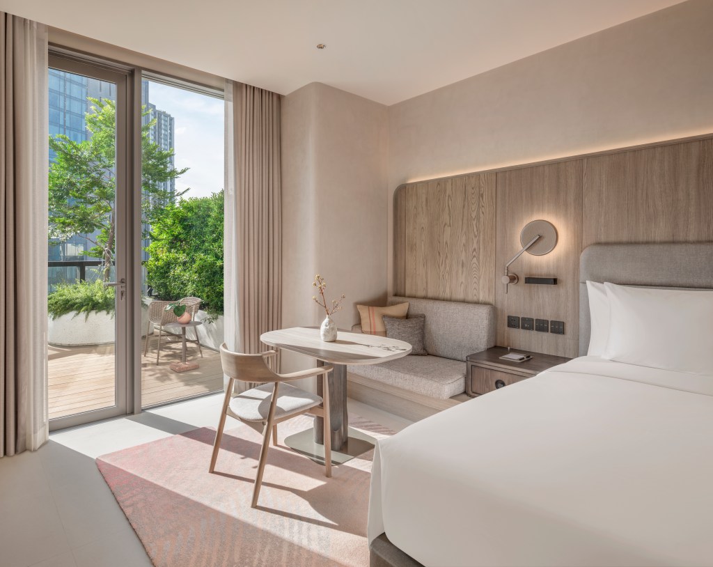 A bedroom with bed, table and chairs, overlooking a baloney with additional chair and greenery.