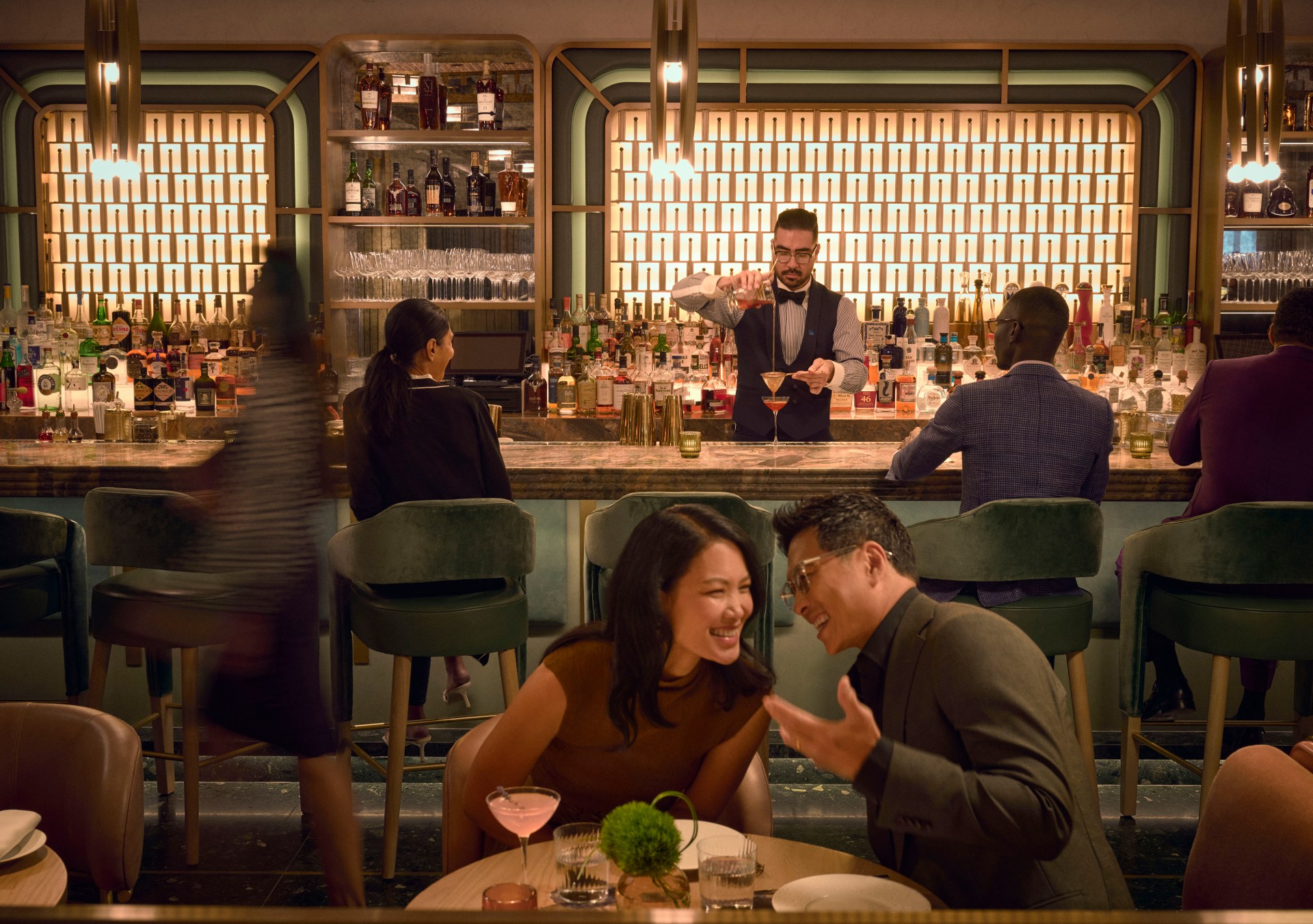 Waldorf Astoria New York - Lex Yard, People sitting on green upholstered barstools and leather chairs in a bar drinking cocktails in coup glassed with a bartender pouring a drink, American brasserie, casually elegant dining room and lively bar, green bar stools