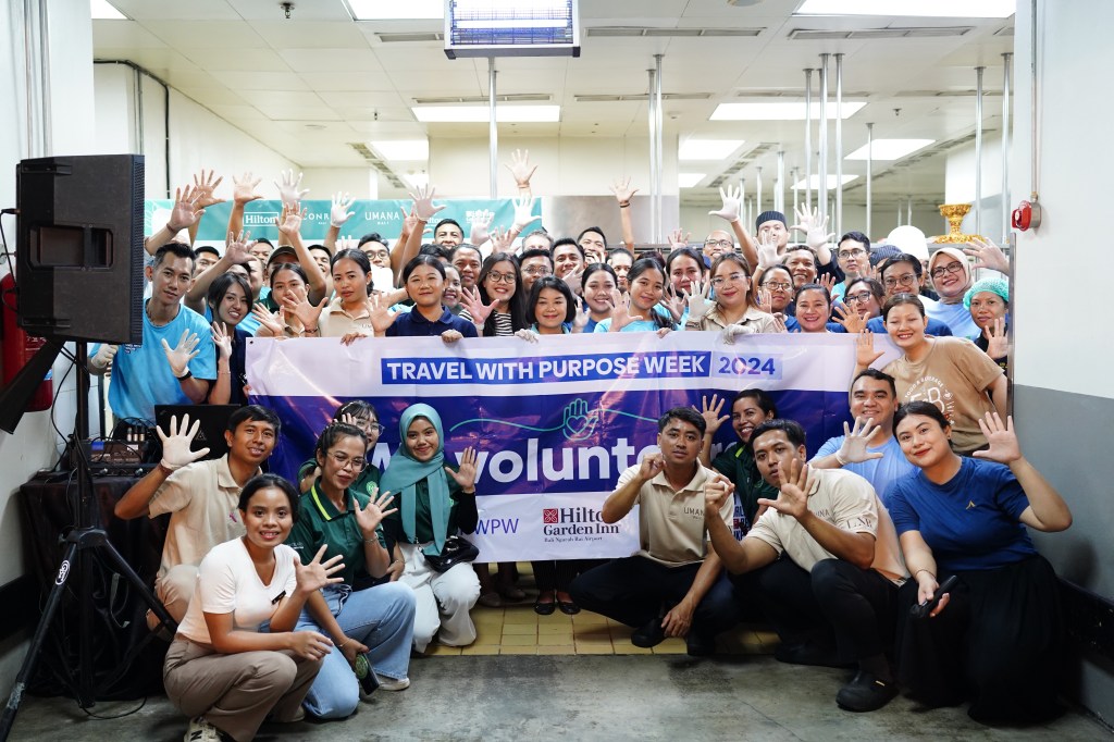 The Hilton Bali team holding up five fingers, smiling at camera with a Travel with Purpose Week banner