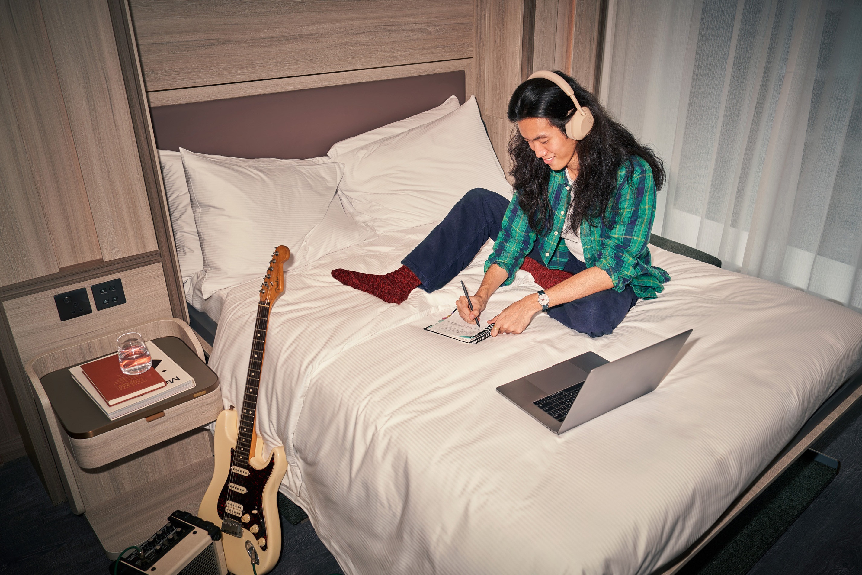 A man sits on the bed making music with guitar and amp next to the bed