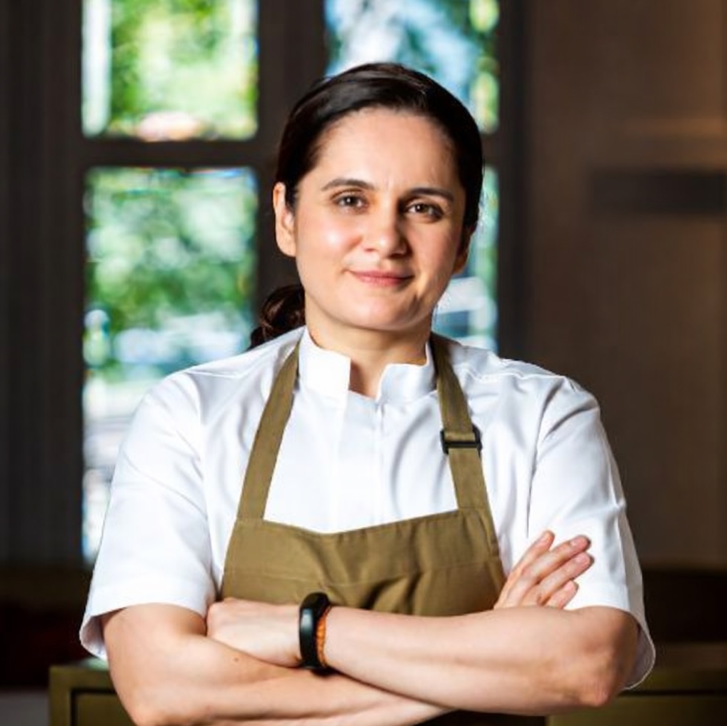 Garima Arora wearing a white chef's jacket and an apron standing with arms crossed in a restaurant setting with a window and greenery in the background.