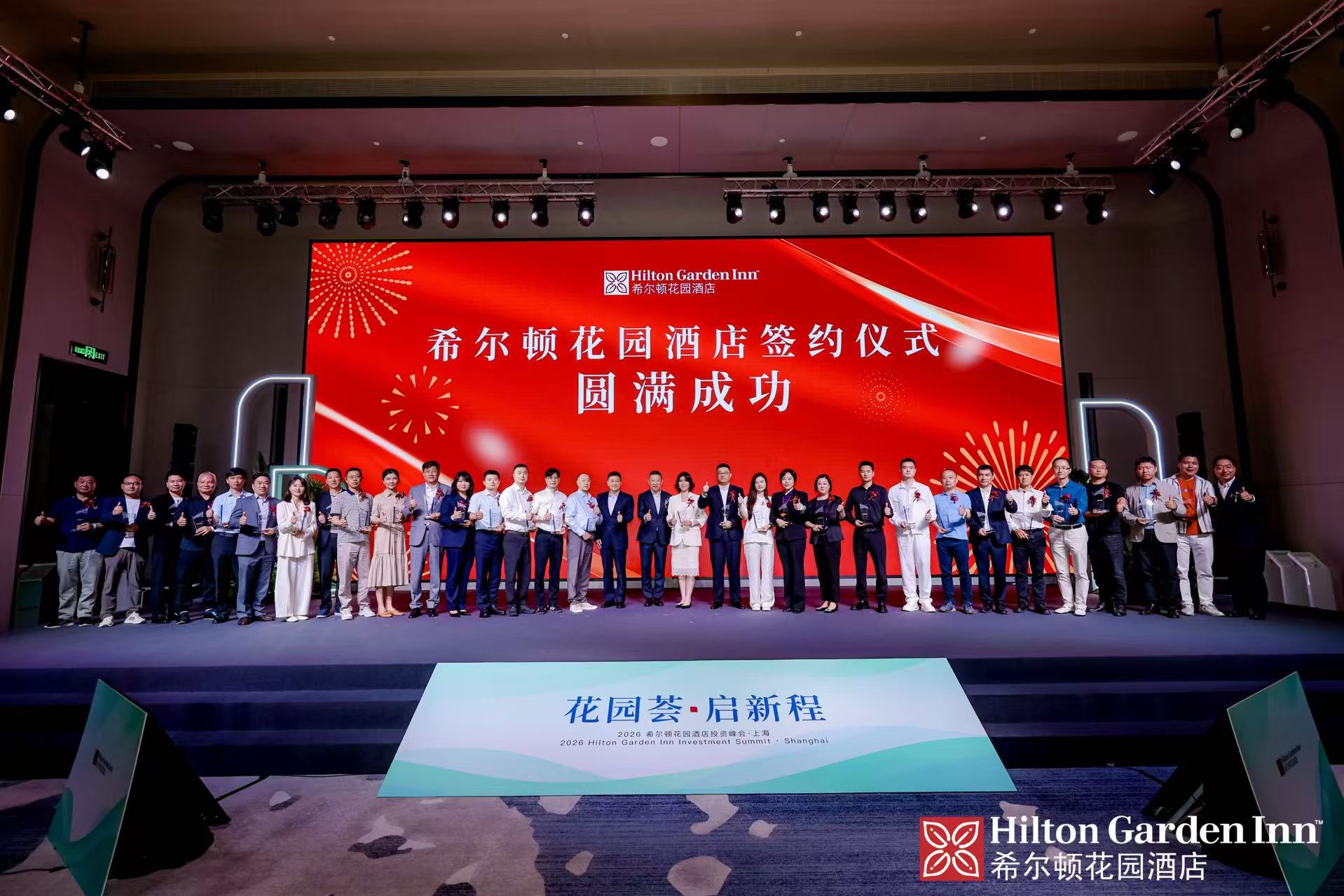 A large group of people standing on a stage in front of a red screen with Hilton Garden Inn branding and Chinese text, celebrating an event.