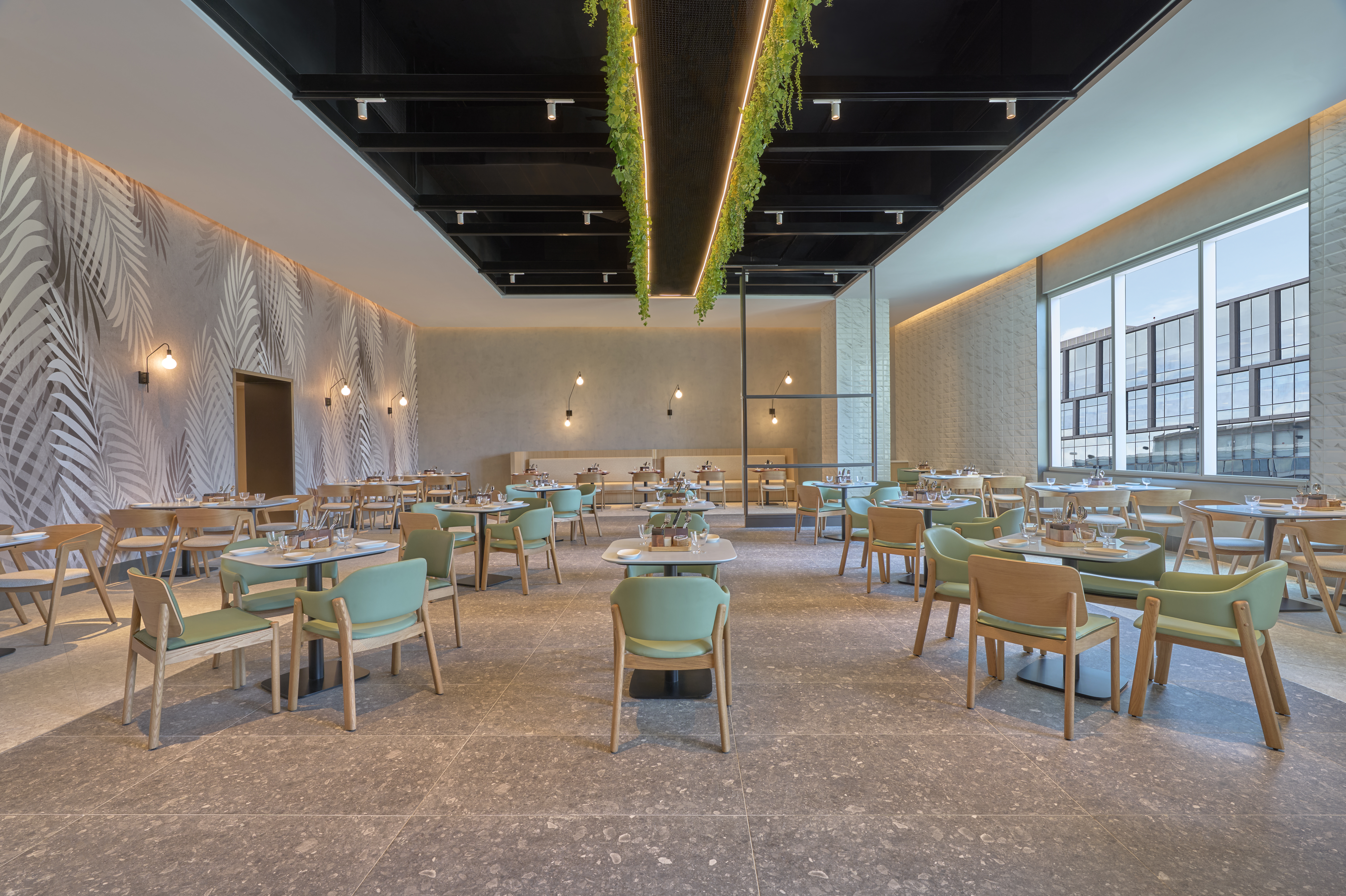 Modern restaurant interior with mint green chairs, wooden tables, large windows, and hanging plants from the ceiling at the Hilton Garden Inn at Embassy TechVillage.