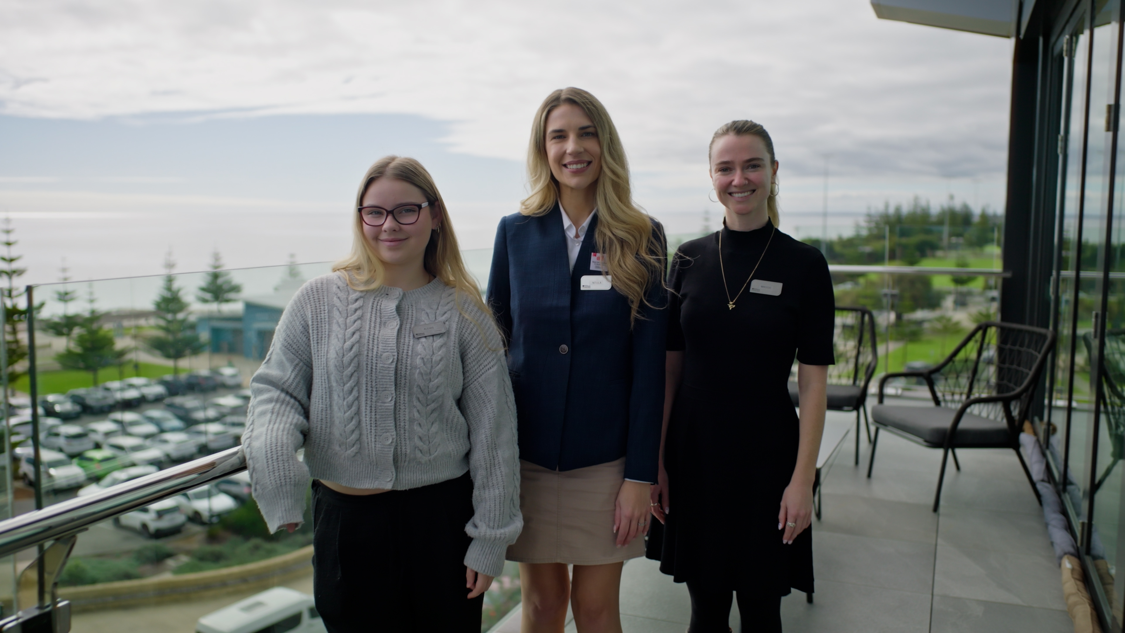 Three Hilton Garden Inn Busselton Team Members standing on a balcony with a parking lot and trees in the background, all smiling and wearing name tags.