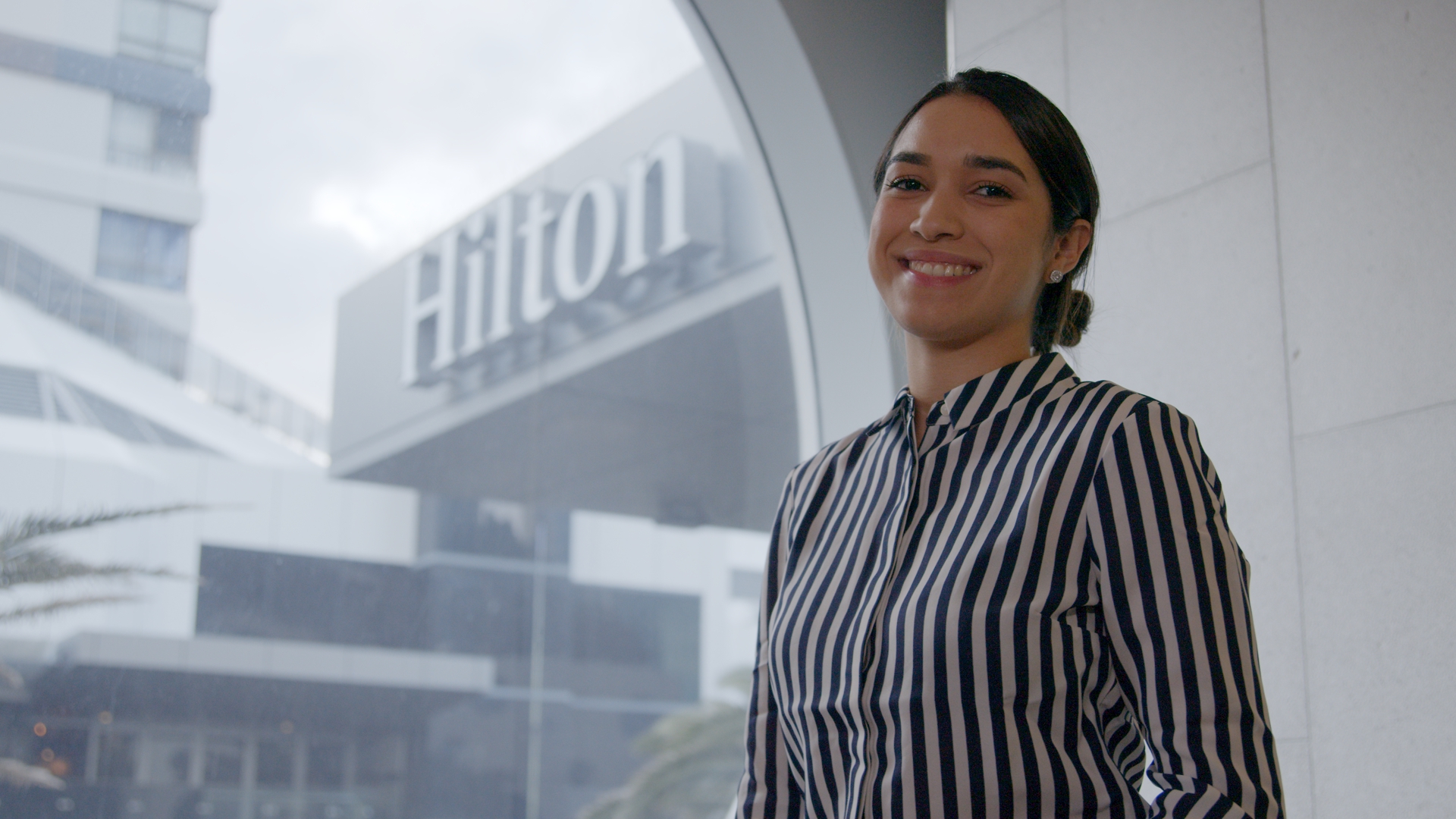 Smiling woman in a striped shirt standing indoors near a window with a blurred Hilton hotel sign visible outside.