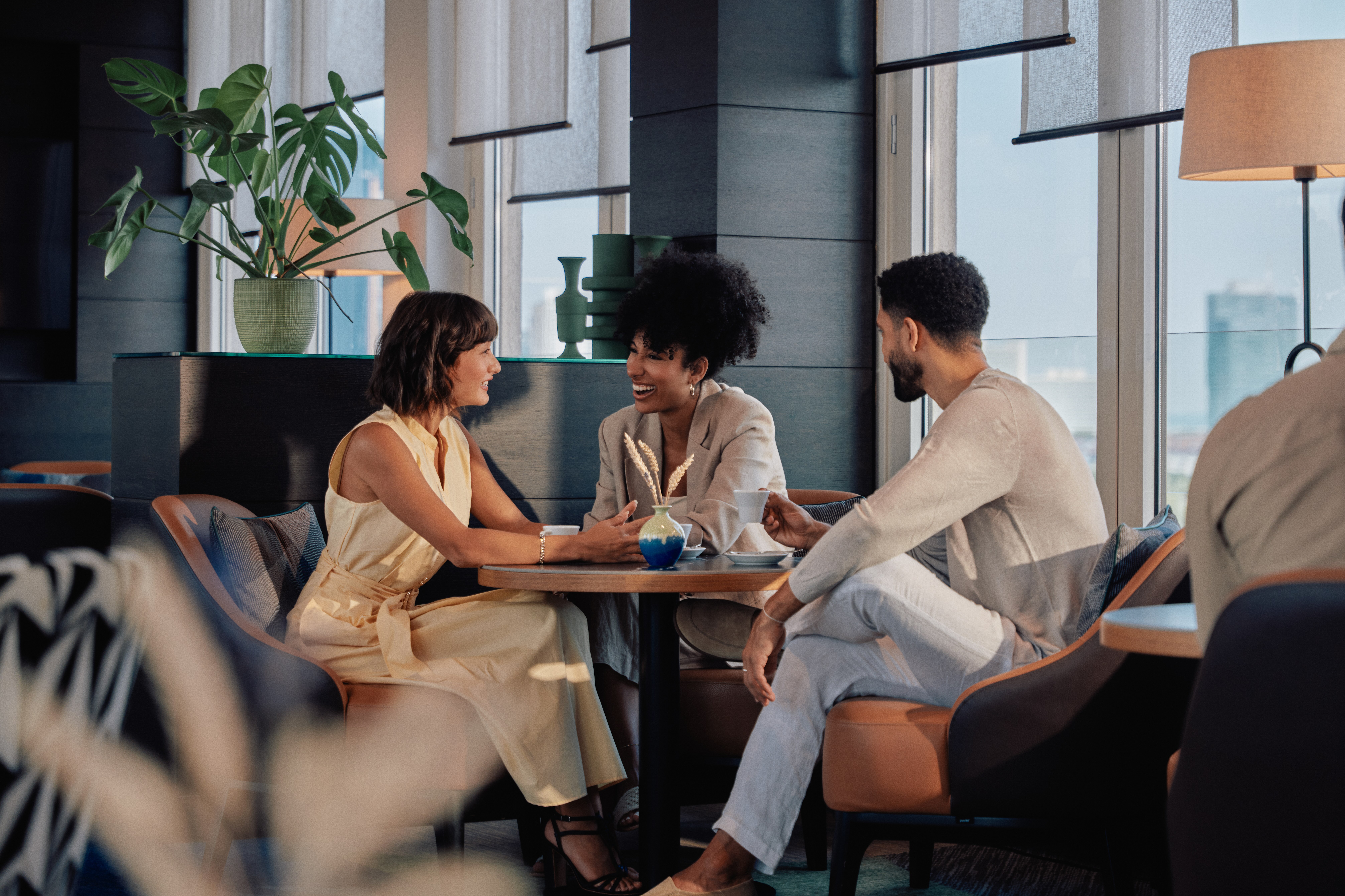 Three people sitting around a table in a modern lounge at Hilton Vienna Waterfront, engaged in a lively conversation with coffee cups in hand.