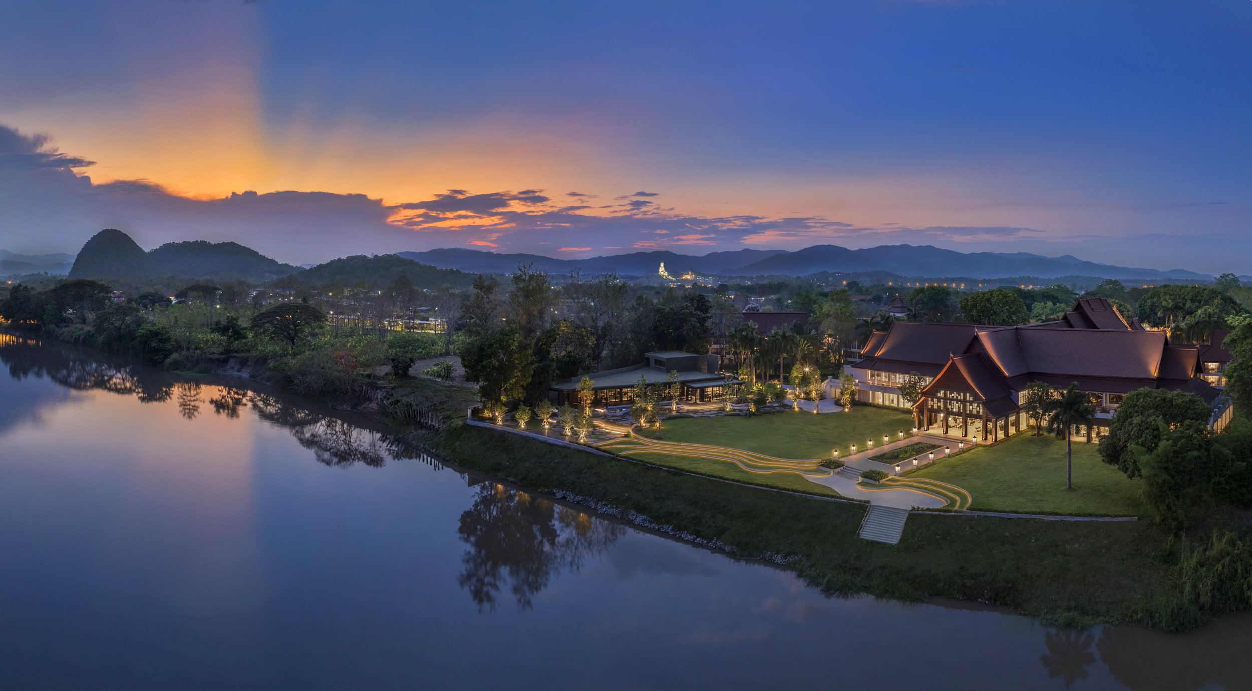 Kahavadi Chiang Rai, Curio Collection by Hilton with traditional architecture lit up at dusk, surrounded by trees and mountains under a colorful sunset sky.