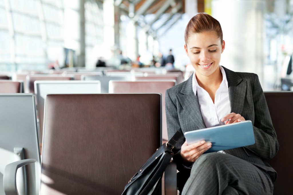 Happy business woman using tablet computer while sitting in departure lounge. Horizontal shot.