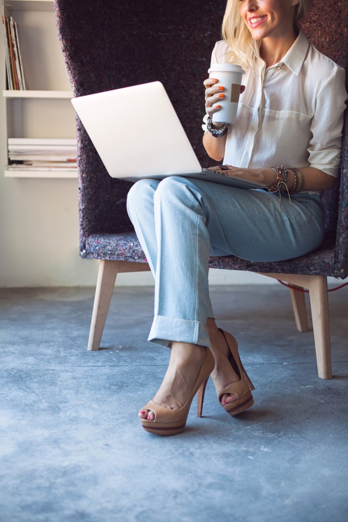 Smiling Caucasian businesswoman sitting in an unusual armchair and working at her laptop while drinking coffee.