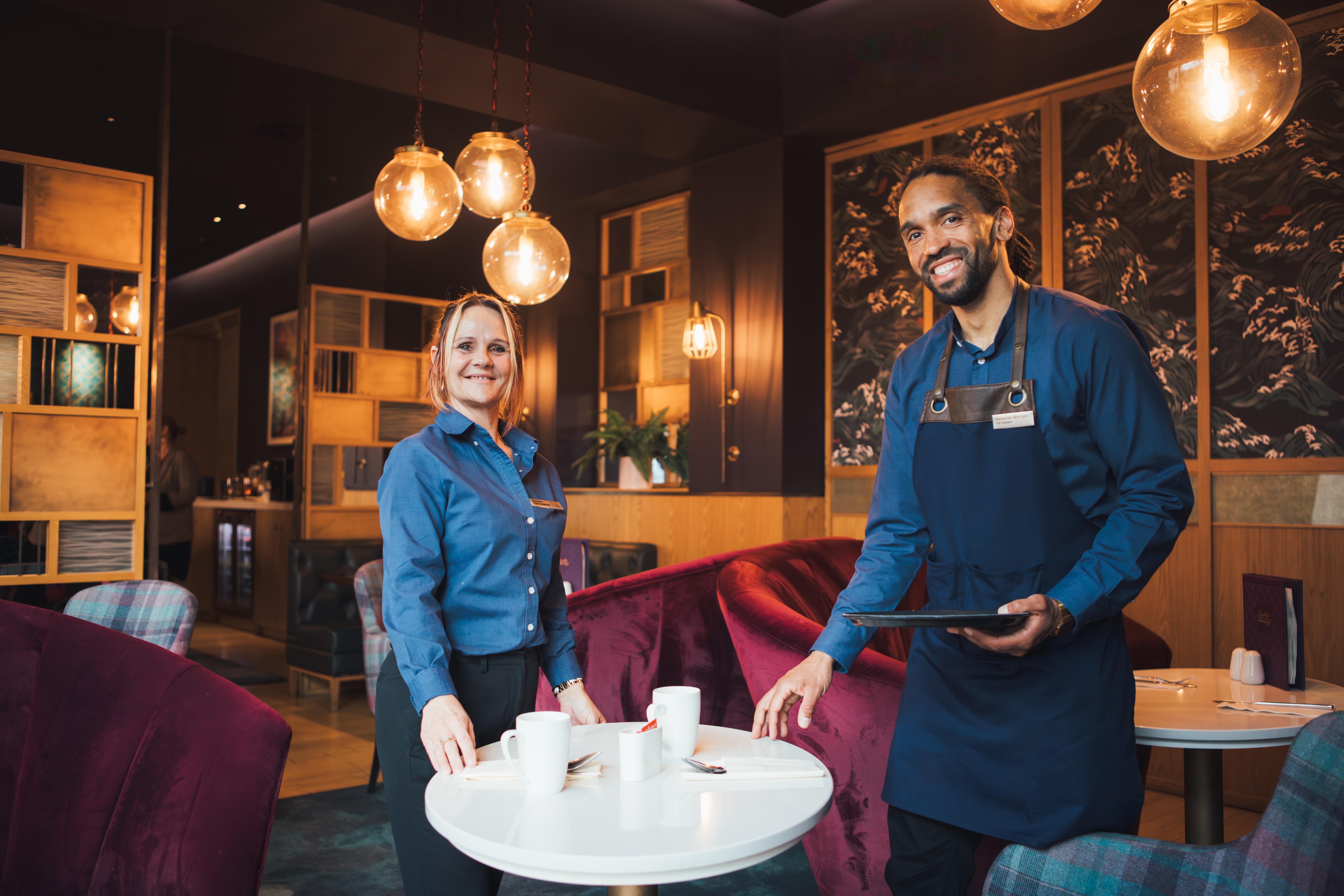 Hilton team members, man and woman standing in a coffee lounge area next to a table with coffee mugs, comfy chairs, small coffee tables, large globe bulb lights, stylistic room partitions in the background