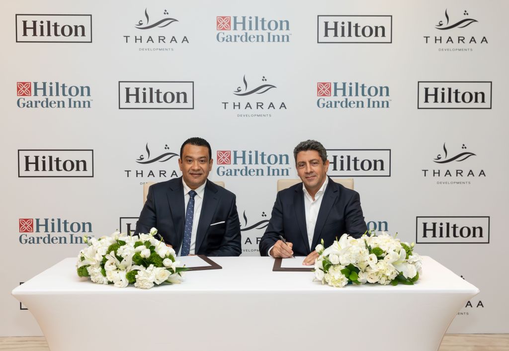 two men sitting at table in front of a back drop with Hilton and Tharaa Development logos, signing agreement