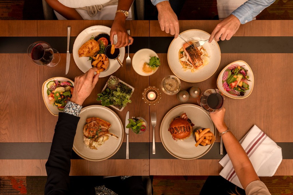 Hilton Woking - OXBO Food - top down view of food on a table people's hands in the frame, eating, fires burgers, fish, wine, salads, porkchops. chicken