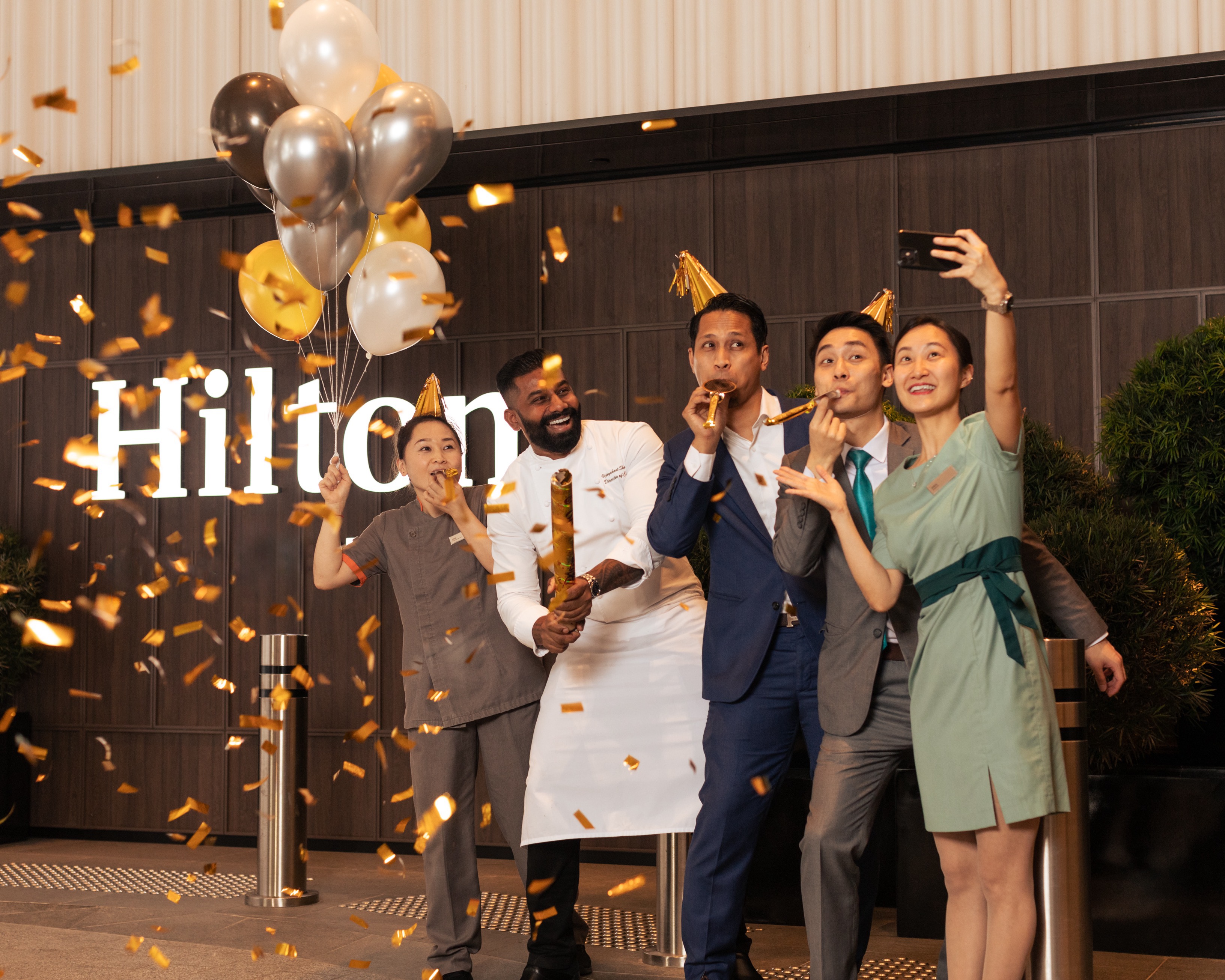 A group of five Hilton Team Members celebrate and take a selfie with balloons, party hats and confetti in front of a Hilton sign