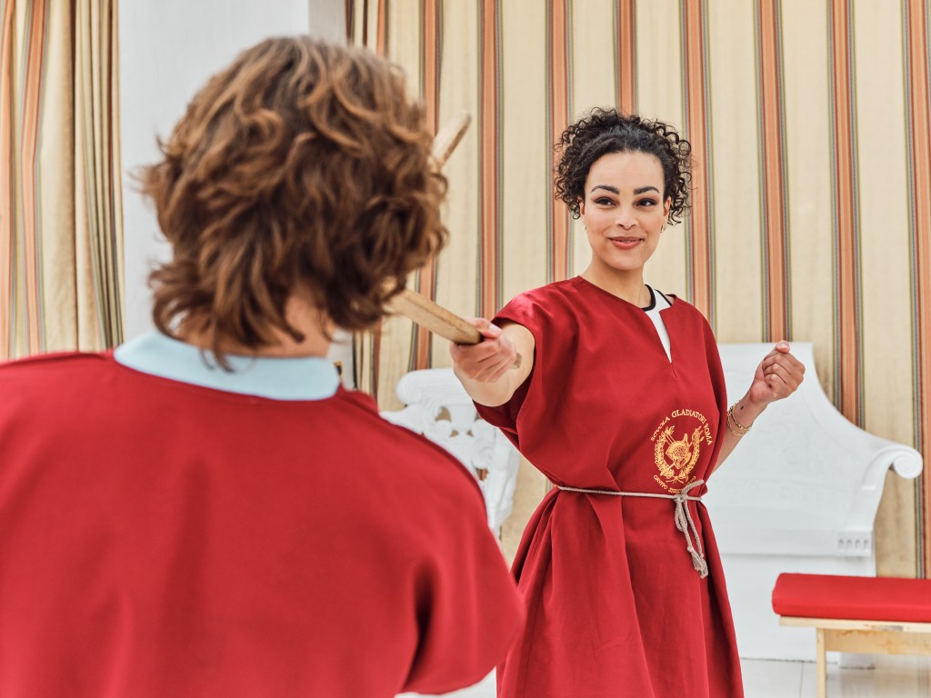 Woman pointing woodedn gladiator sword at man, wearing gladiator training uniform