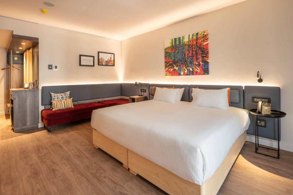 Hampton by Hilton Sandton Grayston - Super King Room, view of bed and side table with phone, art above the bed, couch area with photos above, view of hallway with coffee bar and mini fridge area and clothing rack.