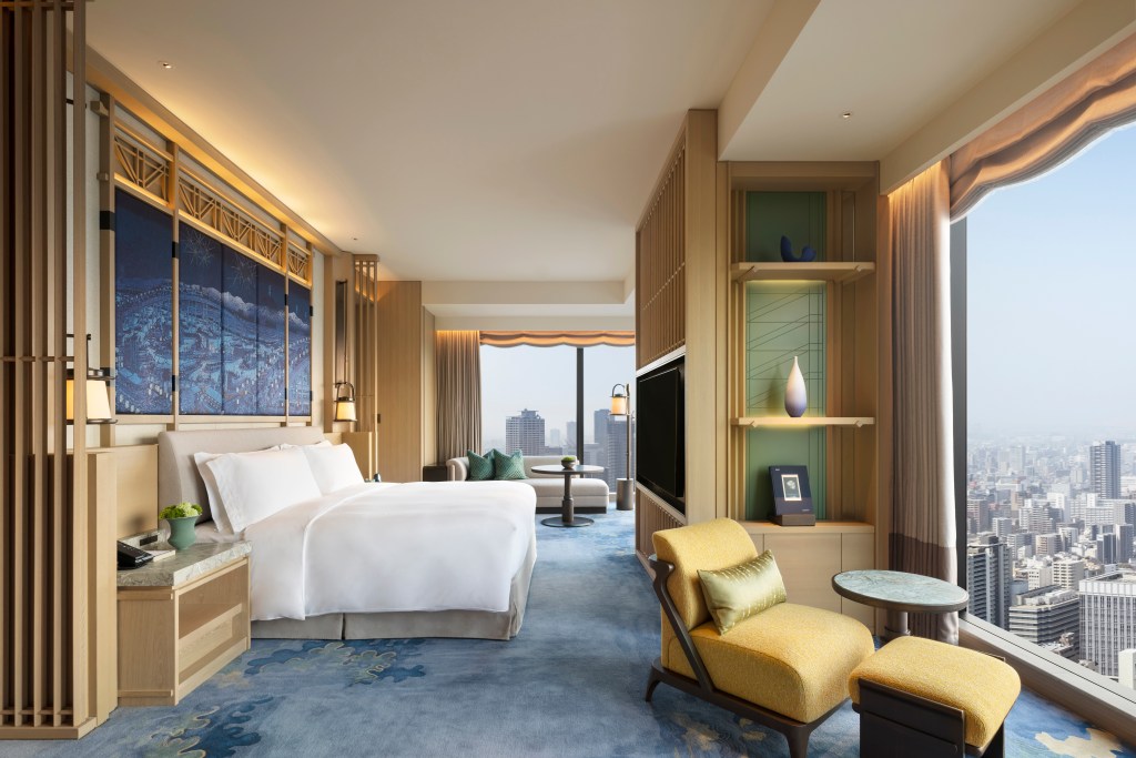 Waldorf Astoria Osaka - Presidential Suite Bedroom, large windows overlooking city skyline, bed with white bedding, large blue city artwork hanging on the wall over bed, table and chair seating area with yellow chair, cream sofa