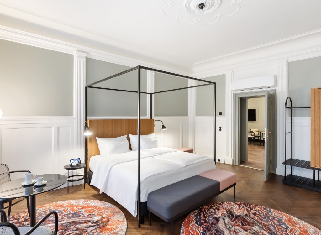 Bedroom with round area rugs with a coffee table with two chairs next to a canopy bed with a wooden headboard and two end tables with lights and framed a framed picture and a wardrobe next to the door
