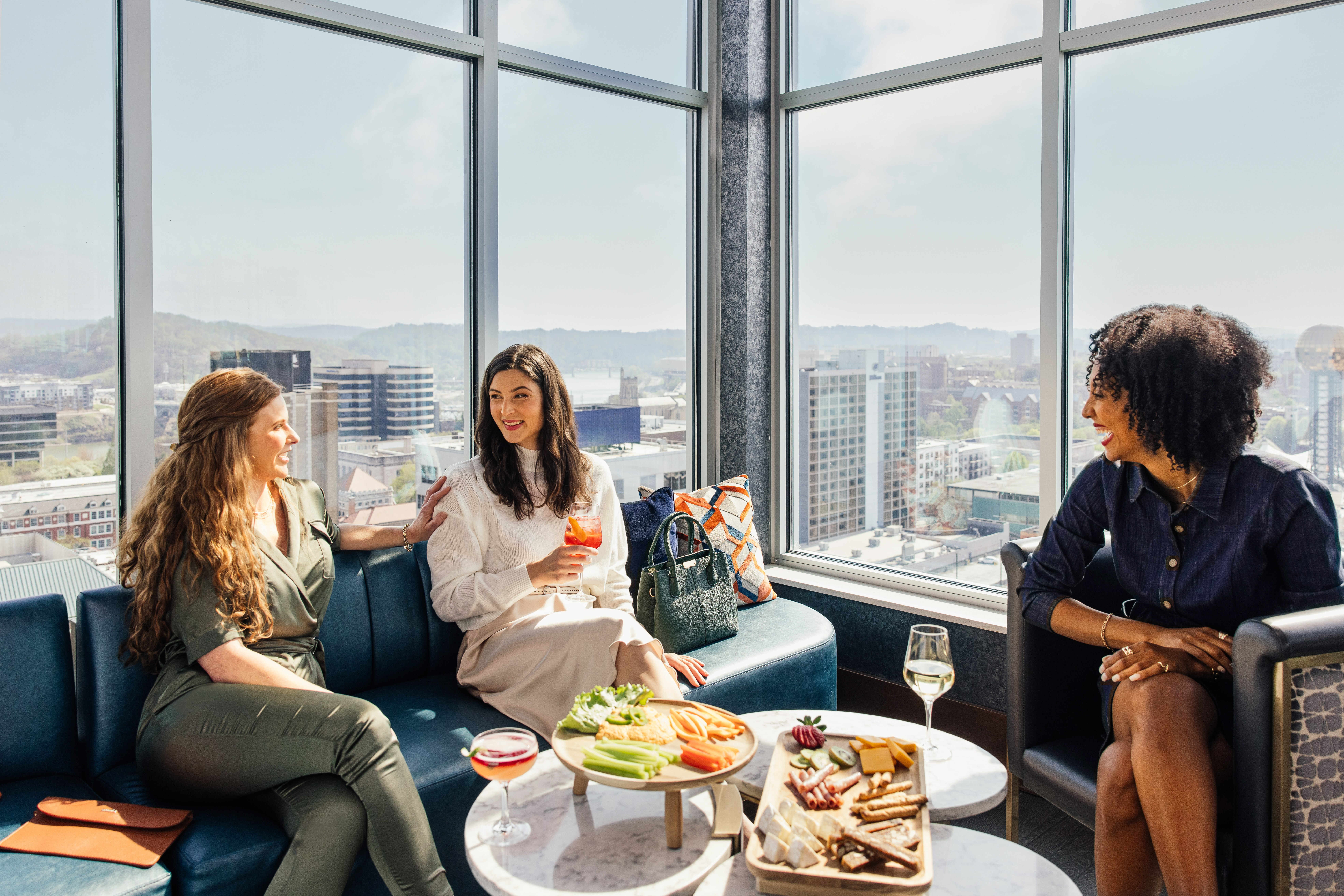 Three women sitting on lounge seats with drinks and appetizers next to windows overlooking the city