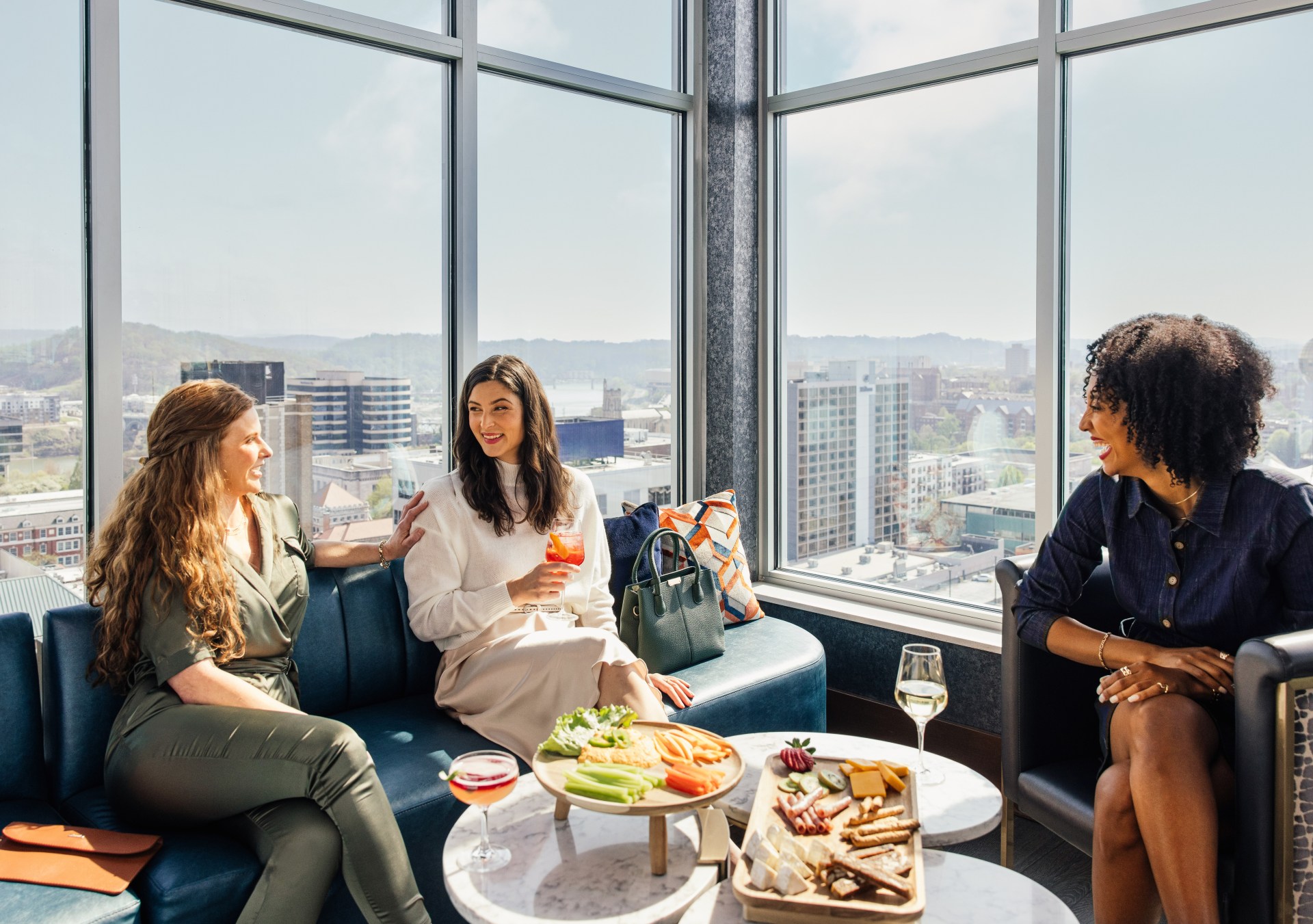 Three women sitting on lounge seats with drinks and appetizers next to windows overlooking the city