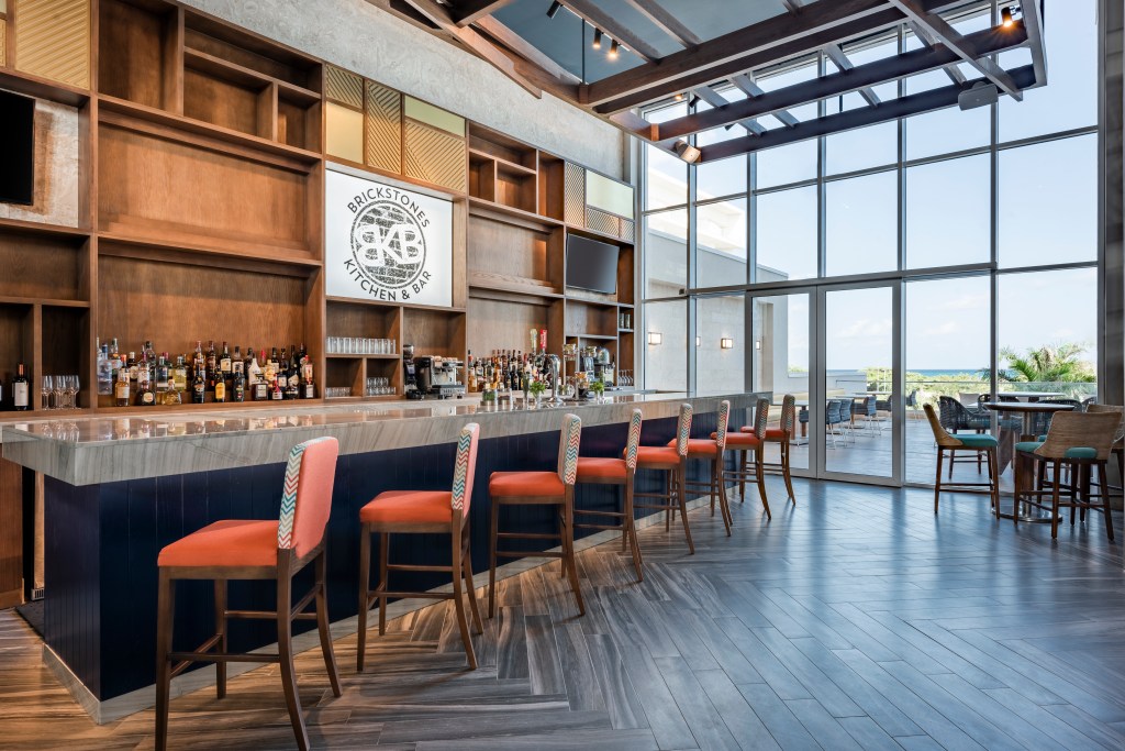 Brickstones Kitchen &amp; Bar at Embassy Suites by Hilton Aruba Resort, Bar with a wooden textured backdrop with colorful bar stools in a room with a floor to ceiling windows and access to outdoor seating overlooking the water