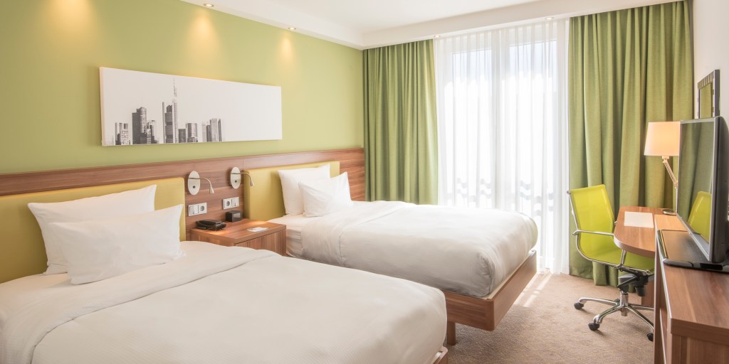 Hampton by Hilton Nuremberg City Centre Twin Guest Room, Twin beds with green and wooden headboard in a room with green walls and curtains along with a green desk chair and artwork hanging on the walls