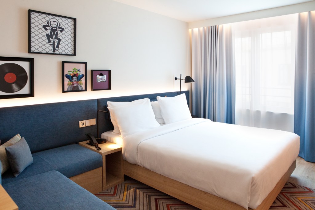 Hampton by Hilton Regensburg Queen Room, Queen bed in a room with blue cushion bench seating along with blue curtains and hanging artwork on the walls