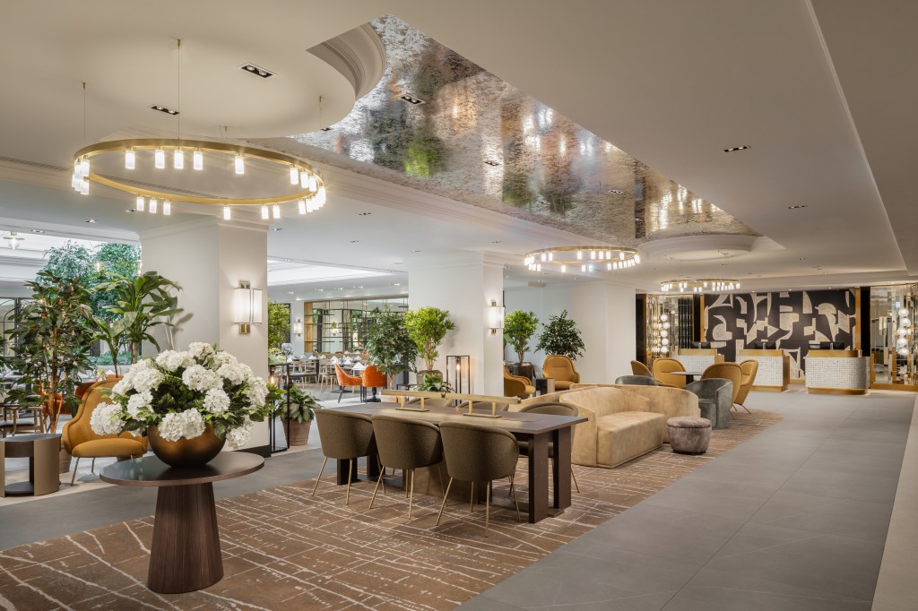 Hilton Heidelberg Lobby, Lobby with flower arrangements on table with round chandelier light above them next to table with chairs for working and more lounge seating for guest with potted plants and a reception desk
